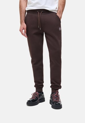 Uomo che indossa pantaloni da jogging marrone scuro con coulisse in vita e stivali neri con lacci, in piedi con una mano in tasca.