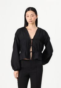 Fekete, rövidített blúz mély V-kivágással és elülső megkötéssel. Puffos ujjakkal és finom redőzött textúrával rendelkezik. Sima anyag.