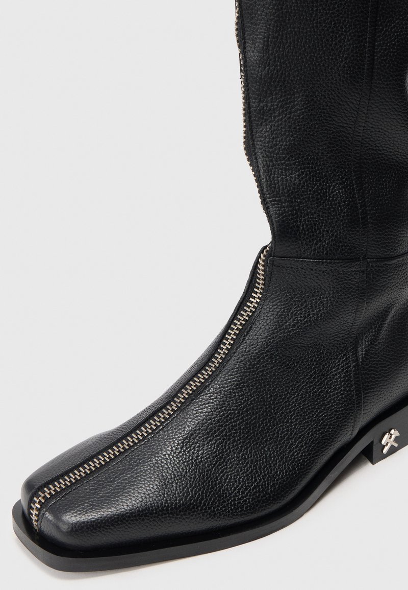 Svart läderstövel med strukturerad yta, fyrkantig tå och en metallisk siddragkedja med silverdetaljer. Tjock svart sula med logotypdetalj.
