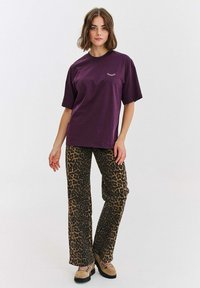 Fialové bavlněné tričko s krátkým rukávem a malou grafikou. Spárováno s hnědými kalhotami s leopardím potiskem a béžovými šněrovacími botami.