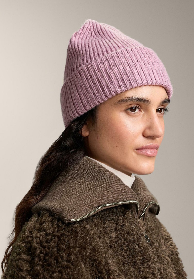 Bonnet rose en tricot côtelé avec un sommet pointu, porté avec une veste texturée marron dotée d'un col zippé. Matières lisses et douces.