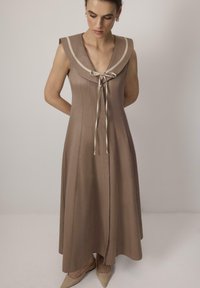 Vrouw in een mouwloze, bruin maxi-jurk met een zeilercollar en strik, gecombineerd met puntige beige ballerina's, staande voor een effen achtergrond.