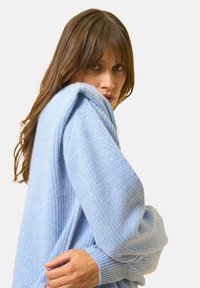 Vaaleansininen neulepaita, jossa on teksturoitu kuvio. Ominaisuuksiin kuuluvat ylisuuret hihansuuhat ja ribbaus. Malli poseeraa yksi käsi vartalon päällä.