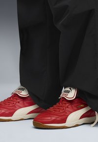 Crvene kožne tenisice s bijelim detaljima i teksturiranim dizajnom. Imaju podstavljeni ovratnik i gumeni potplat. Iznad su vidljive crne hlače.