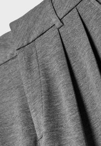 Gros plan d'un pantalon en tissu gris montrant la taille, la boucle de ceinture et les plis avant en matériau doux et texturé.