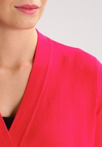 Halenka s výstřihem do V a krátkými rukávy v živé růžové pletenině. Hladká textura s uvolněným střihem, která ukazuje čistý design a minimální detaily.