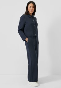 Granatowa krótka kurtka z kołnierzem i zapięciem na guziki z przodu, zestawiona z dopasowanymi spodniami o szerokich nogawkach i białymi trampkami.