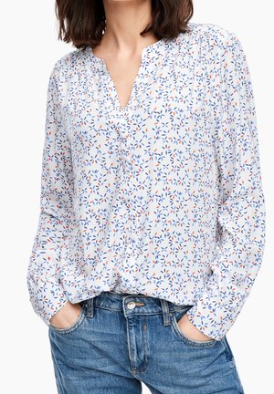 Kobieta ubrana w białą bluzkę z niebiesko-czerwonymi wzorami liści oraz niebieskie dżinsy, z rękami w kieszeniach, na jednolitym białym tle.