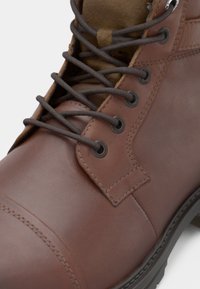 Acercamiento de una bota de cuero marrón con cordones marrones oscuros pasados por ojales de metal y con costuras detalladas.