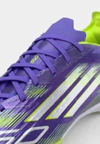 Scarpa sportiva viola con accenti verdi vivaci, caratterizzata da un design elegante, superficie testurizzata e strisce riflettenti argentate. I lacci sono viola.