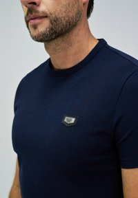 Camiseta de algodón azul marino con cuello redondo y mangas cortas. Presenta un parche de logo rectangular en negro y gris en el lado izquierdo del pecho.