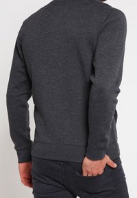 Persona vestida con un suéter de manga larga gris oscuro y pantalones oscuros, de pie con una mano en el bolsillo trasero, vista desde detrás.