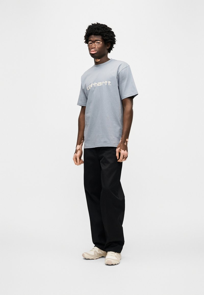 Grijs T-shirt met korte mouwen met "carhartt." in het wit gedrukt, gecombineerd met losse zwarte broek en beige sneakers. Model staat tegen een witte achtergrond.