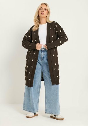 Blondynka w długim ciemnobrązowym kardiganie w białe kropki, białej koszuli, szerokich jasnoniebieskich dżinsach i białych trampkach z czarnymi paskami.