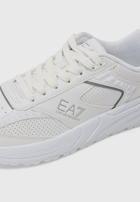 Zapatilla blanca con parte superior de cuero perforado, acentos texturizados y cordones planos. Presenta una marca en el lateral y una suela acolchada.