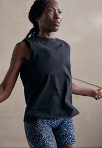 Fekete ujjatlan top, színes, mintás leggings-szel, amely kék és zöld pöttyökkel van díszítve. Az illető ugrókötéllel ugrál benti környezetben.