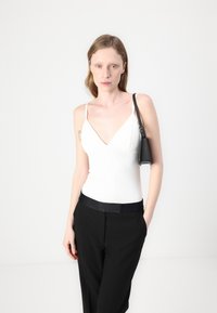 White sleeveless bodysuit with a V-neck design and black high-waisted pants featuring a satin waistband, held with a small black handbag.