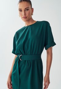 Teal mekko, jossa on lyhyet hihat, vangittu vyötärö hopeisella renkaalla ja sileä, pehmeä tekstuuri.