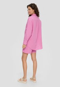 Roze, getextureerde oversized blouse met knoopmanchetten, gecombineerd met bijpassende shorts. Model draagt lichte sandalen, waarbij de benen en het schoeisel duidelijk zichtbaar zijn.