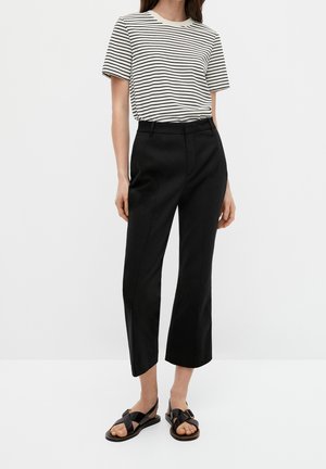 Woman wearing a short-sleeve white shirt with black stripes, black cropped pants, and black open-toe sandals with ankle straps.