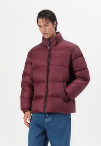 Chaqueta acolchada burdeos con cuello alto, presenta un cierre frontal con cremallera y puños elásticos, combinada con jeans de mezclilla azules.