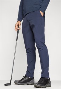 Pantalons de golf bleu marine en tissu léger et extensible ; dotés de deux poches latérales et d'une coupe slim. Associés à des chaussures de golf noires étanches.