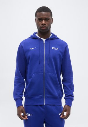 Homme portant un sweat à capuche zippé bleu Nike et un pantalon de survêtement assorti avec le texte « ATLETI », debout devant un fond gris uni.