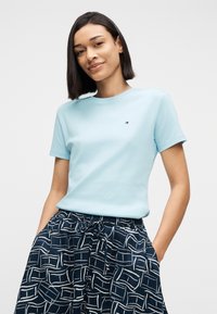 Γυναίκα με σκούρα μαλλιά μέχρι τους ώμους, ντυμένη με ανοιχτό μπλε μπλουζάκι και μπλε σκούρο παντελόνι με σχέδια, στέκεται με τα χέρια στις τσέπες.