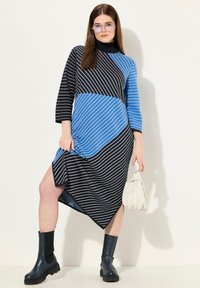Abito a maglia righe in tonalità di nero e blu, con collo alto, maniche a tre quarti, orlo asimmetrico e abbinato a stivali neri alla caviglia.