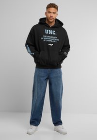 Zwarte hoodie met "UNC" en "University of North Carolina at Chapel Hill" in lichtblauw gedrukt; gecombineerd met lichtblauwe jeans en witte sneakers.