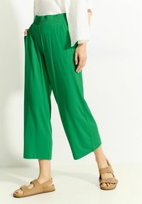 Zielone szerokie spodnie z elastycznym pasem i bocznymi kieszeniami, uszyte z gładkiego materiału. W zestawie z jasnobrązowymi płaskimi sandałami.