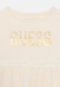 Kreminės spalvos marškiniai su apvalia iškirpte, su auksiniu "GUESS" logotipu, papuoštu blizgančiomis detalėmis, ir tiulio volanu apačioje.