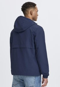 Navy blue hooded jacket with a textured fabric, featuring a back vent, elastic cuffs, and a relaxed fit, paired with light denim.
