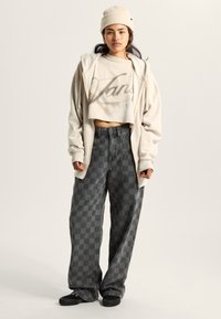 Junge Person trägt eine beige Mütze, einen cremefarbenen Kapuzenpullover mit Reißverschluss, ein kurz geschnittenes T-Shirt mit Grafik, graue karierte Hosen mit weitem Bein und schwarze Slip-On-Schuhe.