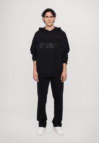 Felpa nera con grafica in rilievo "COUTURE" nera, abbinata a pantaloni neri e sneakers bianche, con un fit rilassato e tasca frontale.