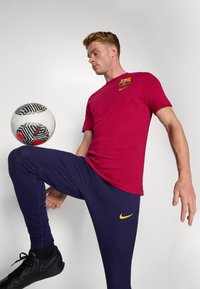 Rød bomulds T-shirt med et Barcelona-logo; marineblå ribbede bukser; sorte sportsko. En fodbold er placeret foran personen.