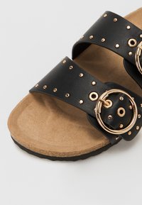 Sandales en simili cuir noir avec deux larges brides ornées de clous dorés et de boucles circulaires, posées sur une semelle texturée couleur tan.