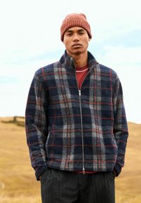 Marineblaue und graue kariert Fleecejacke mit hohem Kragen und Reißverschluss, getragen über einem roten Hemd; ergänzt durch eine gerippte orangefarbene Strickmütze.