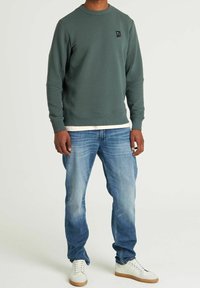 Groene gebreide sweatshirt met ronde hals en geribbelde manchetten, voorzien van een klein zwart logo op de borst, gecombineerd met lichtblauwe spijkerbroek.