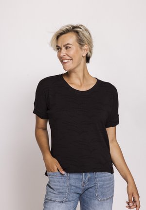 Frau mit kurzen blonden Haaren, die lächelt, trägt ein schwarzes Kurzarmshirt und blaue Denim-Jeans, steht vor einem schlichten hellen Hintergrund.