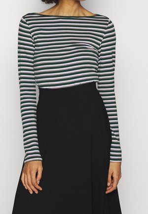 Vrouw draagt een lange mouw top met groene, witte en paarse strepen en een zwarte, hoog opgesneden rok, staand tegen een effen achtergrond.