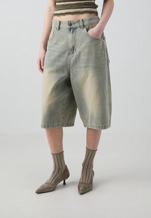 Persoon draagt vervaagde groene lange denimshorts, bruine gestreepte sokken, puntige schoenen met hak en een cropped gestreept topje met één hand in de zak.