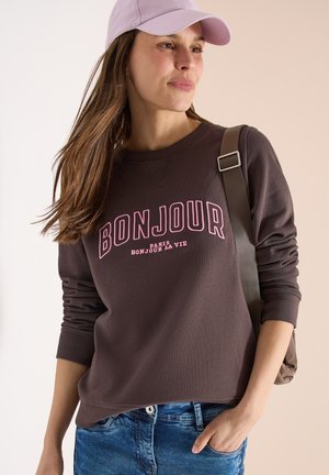 Femme portant une casquette rose, un sweat-shirt marron avec l'inscription "BONJOUR", un jean bleu et un sac à bandoulière, regardant sur le côté avec un léger sourire.