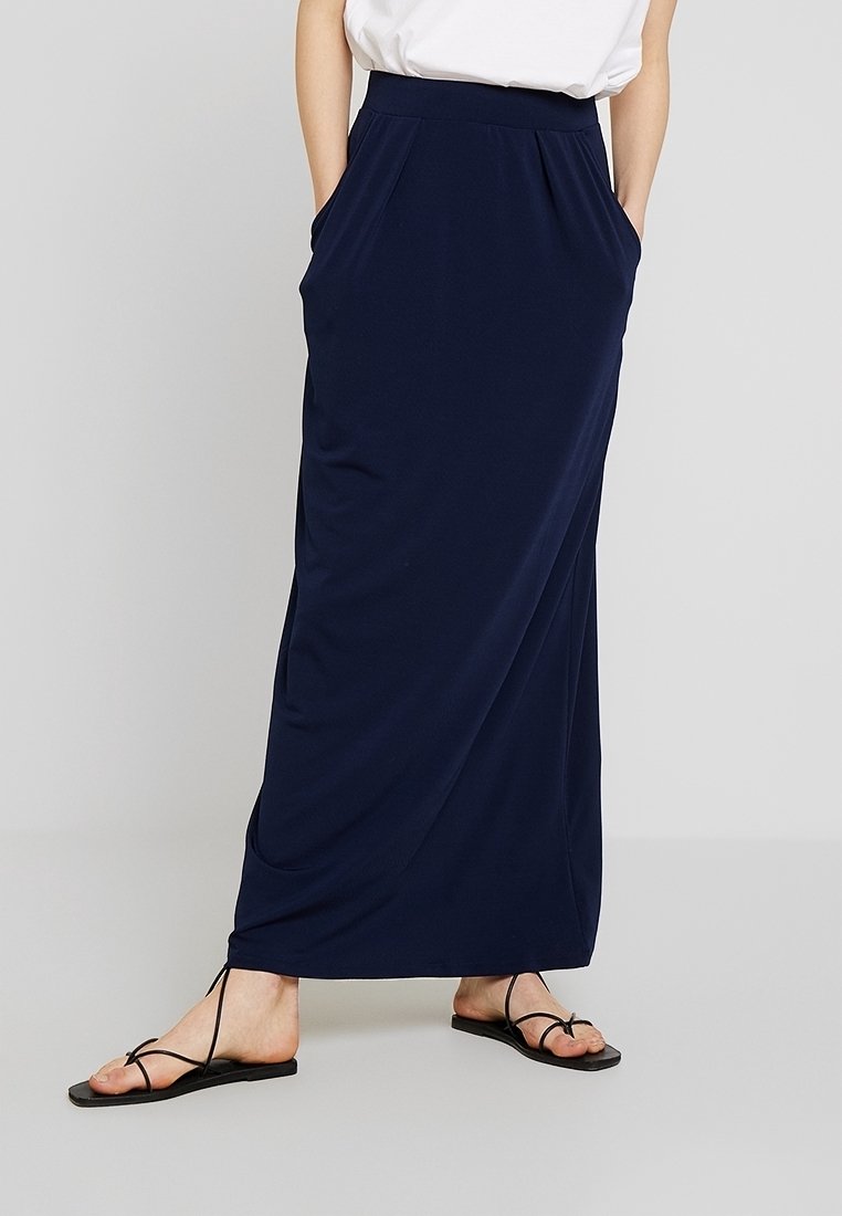 Femme portant une longue jupe bleu marine avec des poches et des sandales plates noires à lanières, debout devant un fond uni.