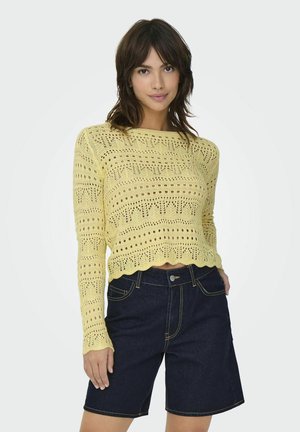 Suéter corto de punto amarillo con patrones intrincados y dobladillo con ondas, combinado con pantalones cortos de mezclilla oscura con costuras en contraste.