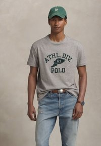 Joven vestido con una camiseta gris que dice "ATHL. DIV. POLO", gorra verde, jeans azul claro, cinturón de cuero marrón y un reloj en su muñeca izquierda.