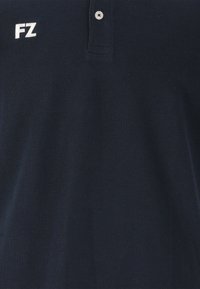Mörkblå polo skorta i strukturerad tyg, med en treknappsknäppning och de vita broderade bokstäverna "FZ" på vänster bröst.