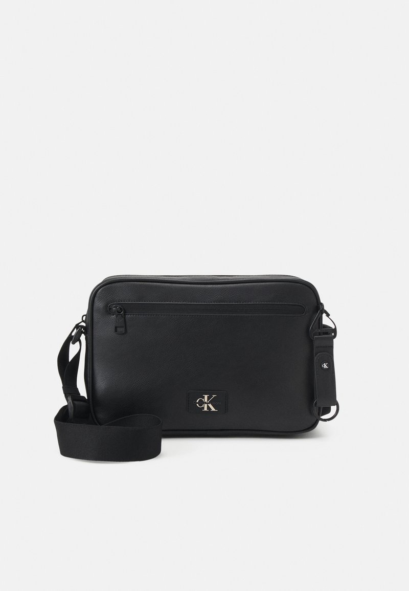 Musta suorakulmainen olkalaukku, jossa on säädettävä hihna, etupuolella vetoketjutasku ja pieni Calvin Klein -logo.