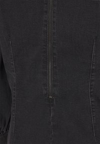 Sort denimstof med en centreret lodret metallynlås og synlige syninger langs sømmene i et close-up.