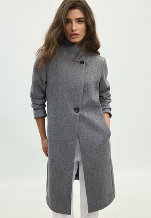 Femme aux longs cheveux bruns portant un manteau en laine gris avec deux boutons noirs, debout avec une main dans la poche sur un fond clair.
